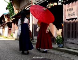 Two women in traditional hakama and kimono, one holding a large red parasol, walking down a preserved street in Mikuni Minato.