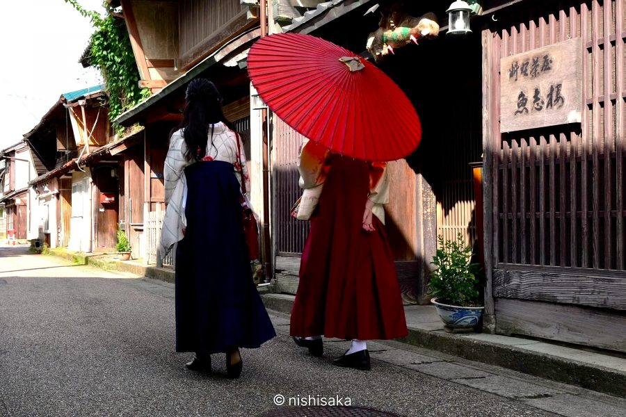 Two women in traditional hakama and kimono, one holding a large red parasol, walking down a preserved street in Mikuni Minato.
