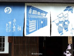 Three blue and white printed noren curtains displaying traditional town illustrations and Japanese text, hanging in front of a wooden shop facade.