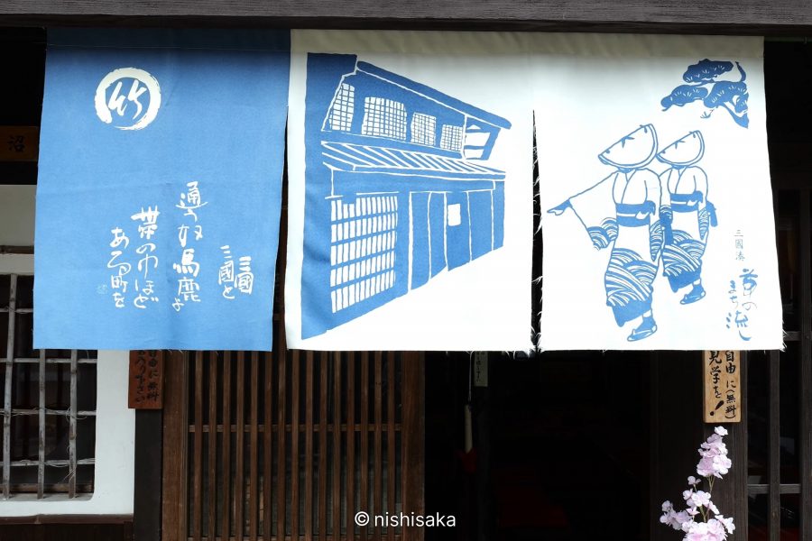 Three blue and white printed noren curtains displaying traditional town illustrations and Japanese text, hanging in front of a wooden shop facade.