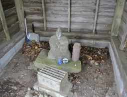 A weathered stone Jizo statue housed in a small wooden shelter, with offerings placed at its base.