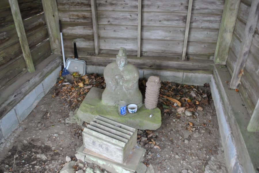 A weathered stone Jizo statue housed in a small wooden shelter, with offerings placed at its base.