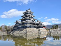 The main keep of Matsumoto Castle reflected in the surrounding moat under a bright blue sky with scattered clouds.