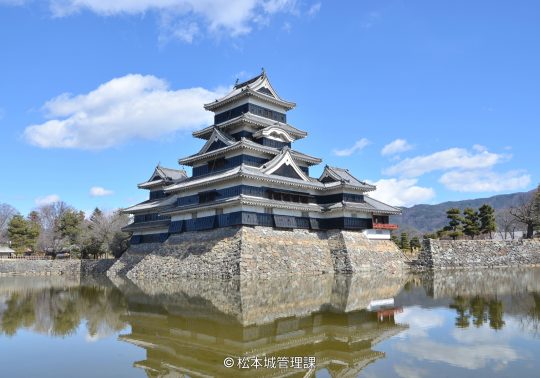 The main keep of Matsumoto Castle reflected in the surrounding moat under a bright blue sky with scattered clouds.