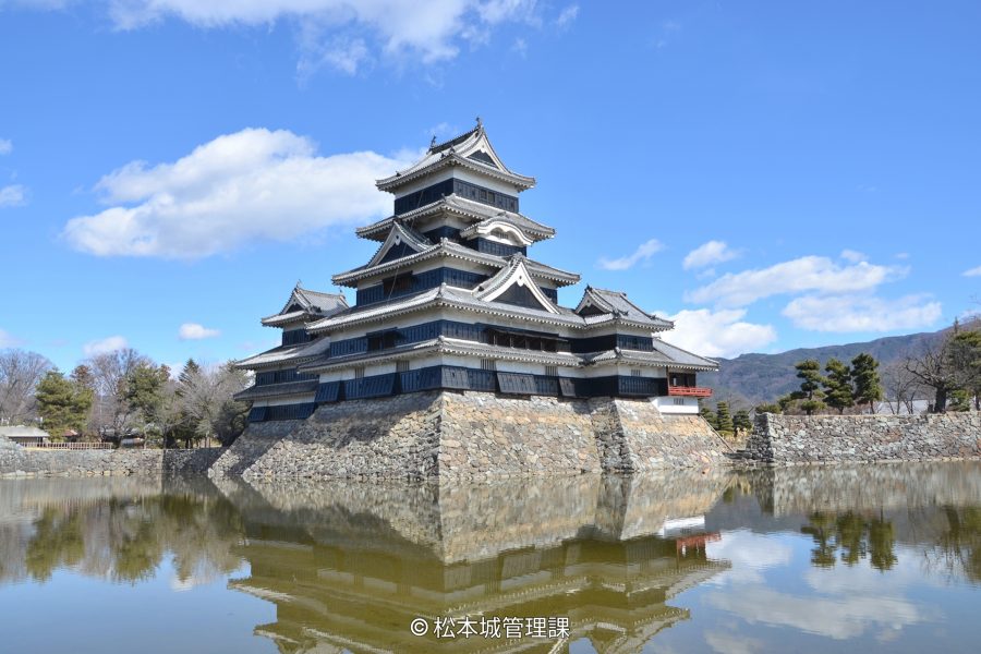The main keep of Matsumoto Castle reflected in the surrounding moat under a bright blue sky with scattered clouds.