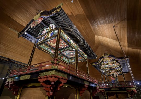 Two ornate, large Japanese festival floats (dashi or yatai) with detailed carvings and painted roofs, displayed in a modern wooden museum room.
