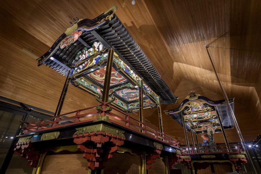 Two ornate, large Japanese festival floats (dashi or yatai) with detailed carvings and painted roofs, displayed in a modern wooden museum room.