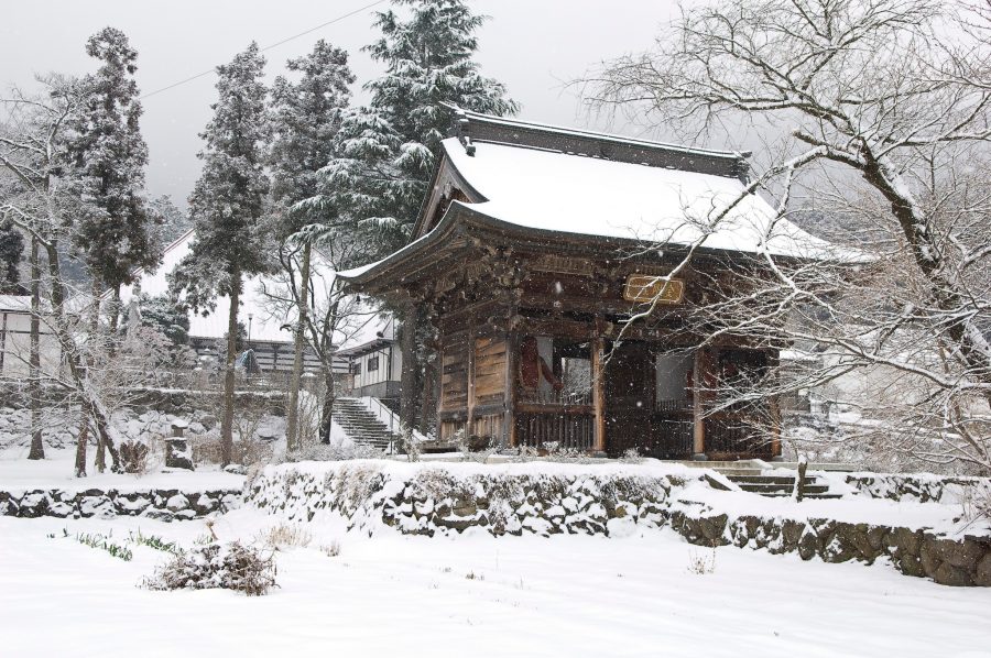 A traditional Japanese temple building covered in snow, surrounded by trees and a snow-covered landscape on a winter day.