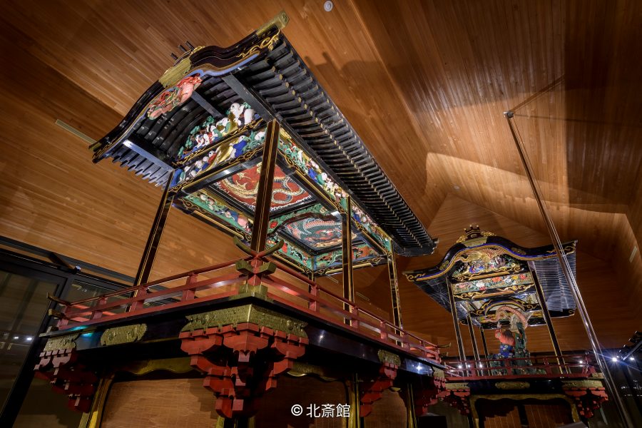 Two ornate, large Japanese festival floats (dashi or yatai) with detailed carvings and painted roofs, displayed in a modern wooden museum room.