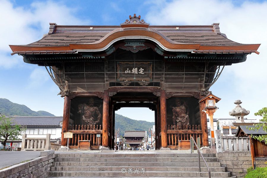 The majestic two-story Sanmon Gate of Zenko-ji Temple, featuring carved Nio statues in alcoves, viewed from stone steps leading up to the main street.