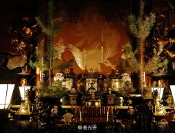A close-up, dim view of the ornate Buddhist altar inside a temple hall, featuring golden lanterns, tall pine branches, and a gold phoenix embroidery on the dark red backdrop.