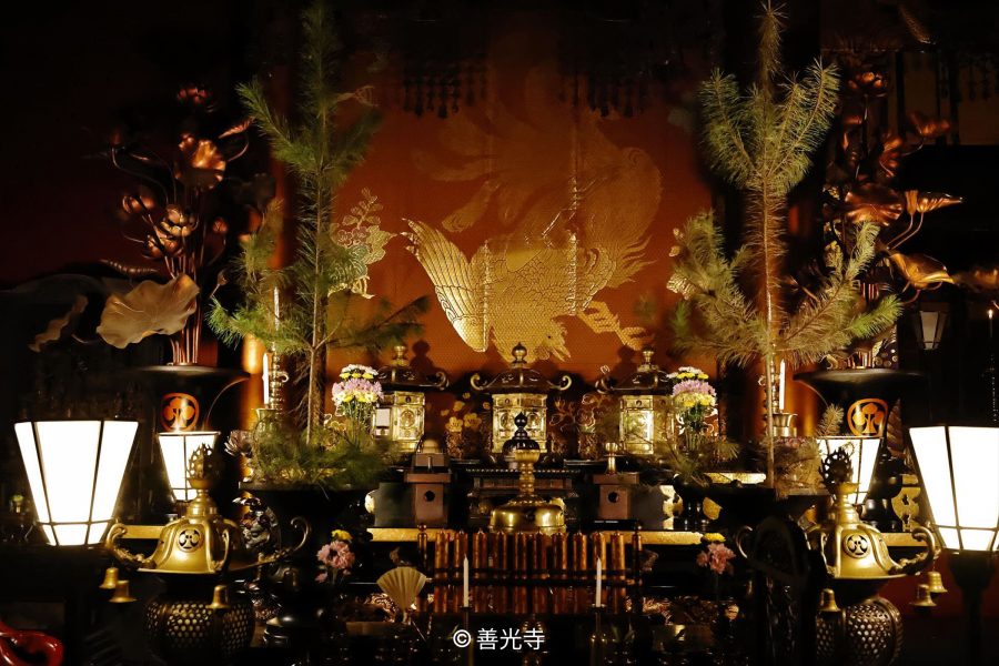 A close-up, dim view of the ornate Buddhist altar inside a temple hall, featuring golden lanterns, tall pine branches, and a gold phoenix embroidery on the dark red backdrop.