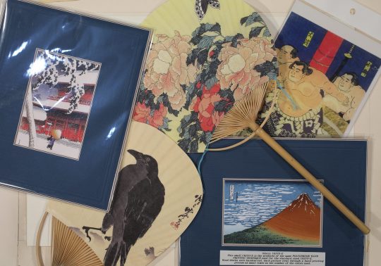 A close-up of various ukiyo-e-style art objects, including framed prints of a snow scene and Mount Fuji, and traditional folding fans (uchiwa) featuring a crow, flowers, and sumo wrestlers.