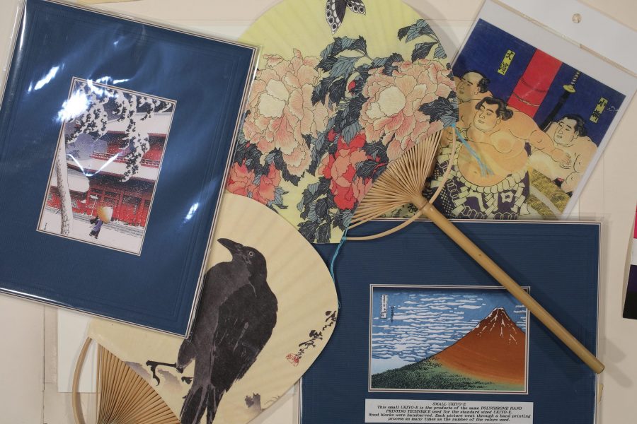 A close-up of various ukiyo-e-style art objects, including framed prints of a snow scene and Mount Fuji, and traditional folding fans (uchiwa) featuring a crow, flowers, and sumo wrestlers.