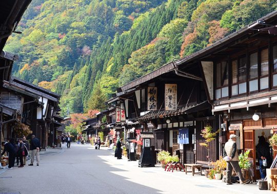 A wide street view of Narai-juku in autumn, with people walking along the preserved wooden shops and green and yellow foliage covering the surrounding mountains.