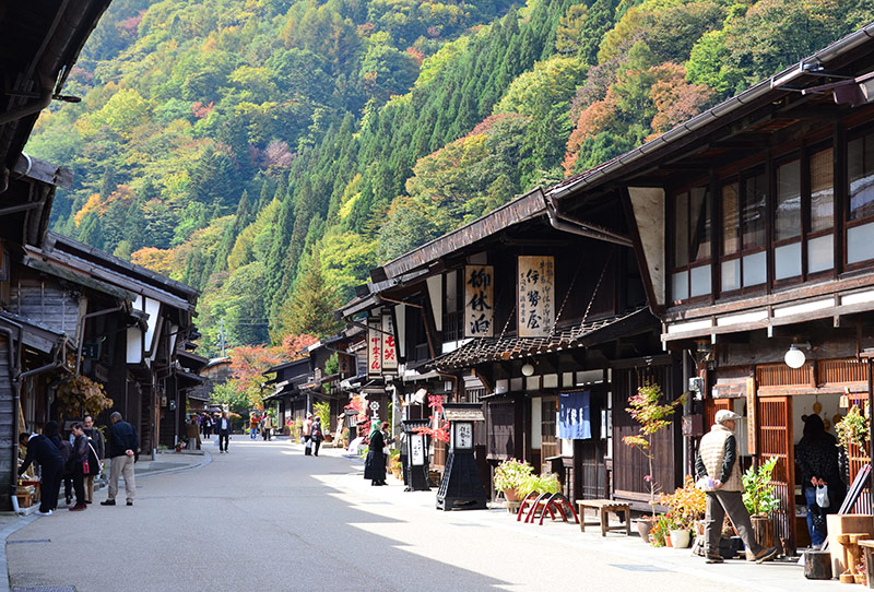 A wide street view of Narai-juku in autumn, with people walking along the preserved wooden shops and green and yellow foliage covering the surrounding mountains.