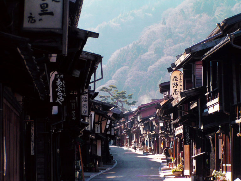 A narrow, gently curving street lined with traditional dark wooden buildings of the post town, leading up towards a mist-shrouded, tree-covered mountain slope.