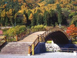 A highly arched, traditional wooden drum bridge (Taiko-bashi) over water, surrounded by vibrant autumn trees in yellow, orange, and green.