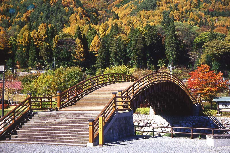 A highly arched, traditional wooden drum bridge (Taiko-bashi) over water, surrounded by vibrant autumn trees in yellow, orange, and green.