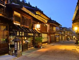 Narai-juku street at dusk, illuminated by warm street lights, showing preserved Edo-period wooden buildings with intricate latticework.