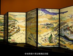 A multi-panel screen painting (byobu) with a detailed depiction of a historic battle or procession on a gold background.