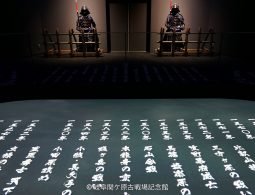 A dark, theatrical museum exhibit showing two samurai armor sets and illuminated Japanese text (historical dates and names) projected onto the floor.