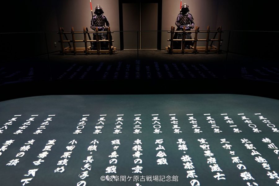 A dark, theatrical museum exhibit showing two samurai armor sets and illuminated Japanese text (historical dates and names) projected onto the floor.