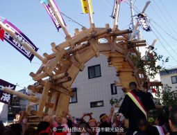 A large, complex wooden structure (likely a parade float or portable shrine) being carried above a crowd during a festival in a town setting.