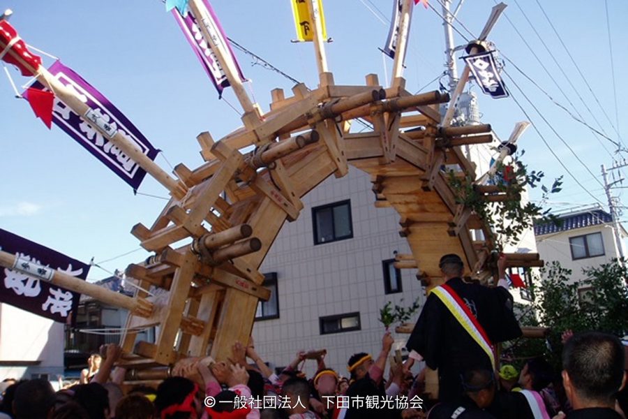A large, complex wooden structure (likely a parade float or portable shrine) being carried above a crowd during a festival in a town setting.