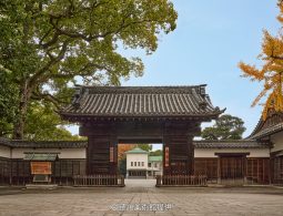 A traditional black wooden gate and white earthen wall, leading to a courtyard with a secondary building in the distance.