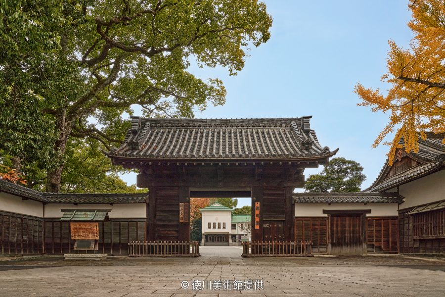 A traditional black wooden gate and white earthen wall, leading to a courtyard with a secondary building in the distance.