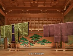 An exhibition room display of two ornate silk kimonos (likely Noh costumes or ceremonial robes) in front of a painted pine tree backdrop.