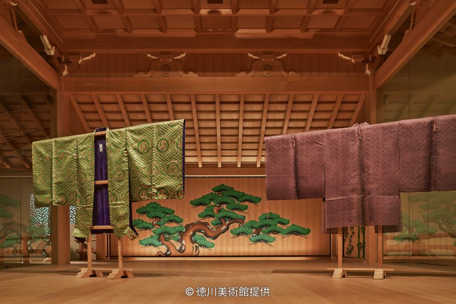An exhibition room display of two ornate silk kimonos (likely Noh costumes or ceremonial robes) in front of a painted pine tree backdrop.