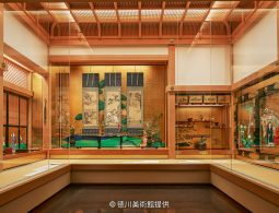 A museum room resembling a traditional Japanese palace chamber, displaying screens, hanging scrolls, and lacquerware in glass cases.