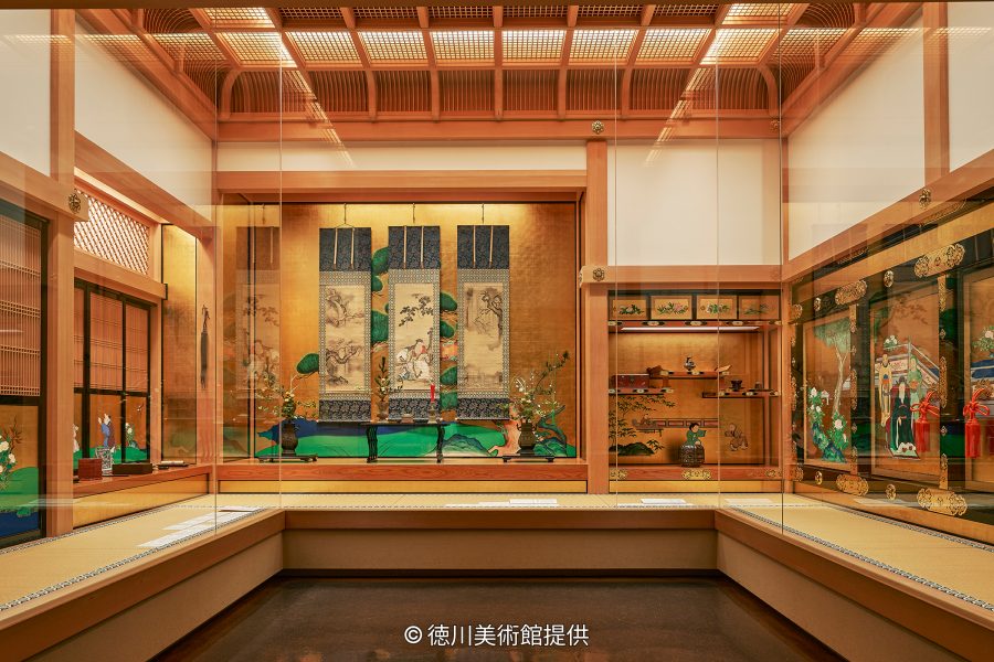 A museum room resembling a traditional Japanese palace chamber, displaying screens, hanging scrolls, and lacquerware in glass cases.