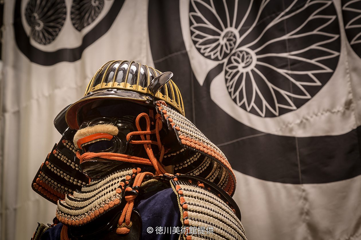 Close-up of a samurai helmet (kabuto) and face guard (menpo) armor, displayed in front of a banner with the Tokugawa family crest.