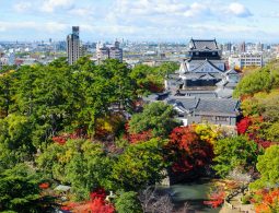 An elevated view of Okazaki Castle keep and surrounding park, showing a mixture of green, red, and yellow autumn foliage, with the modern city in the background.