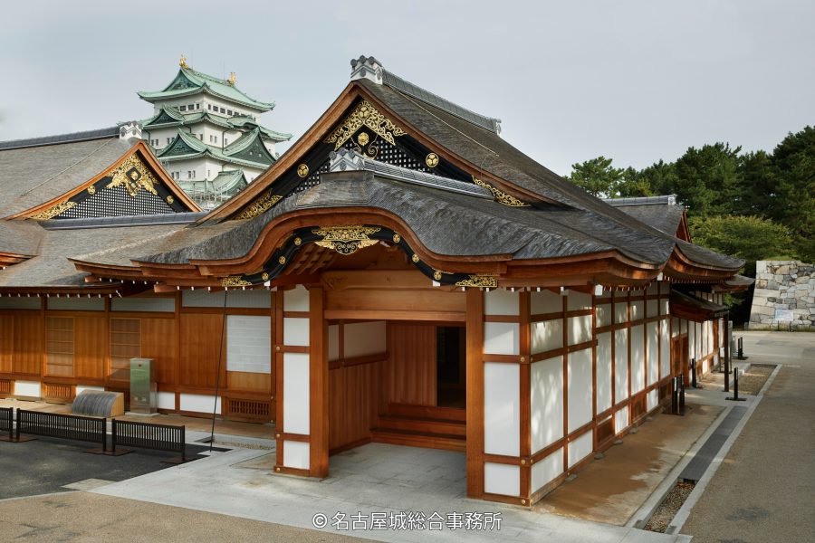The entrance building of the reconstructed Honmaru Palace, showing intricate eaves and woodwork, with the main castle keep visible in the background.