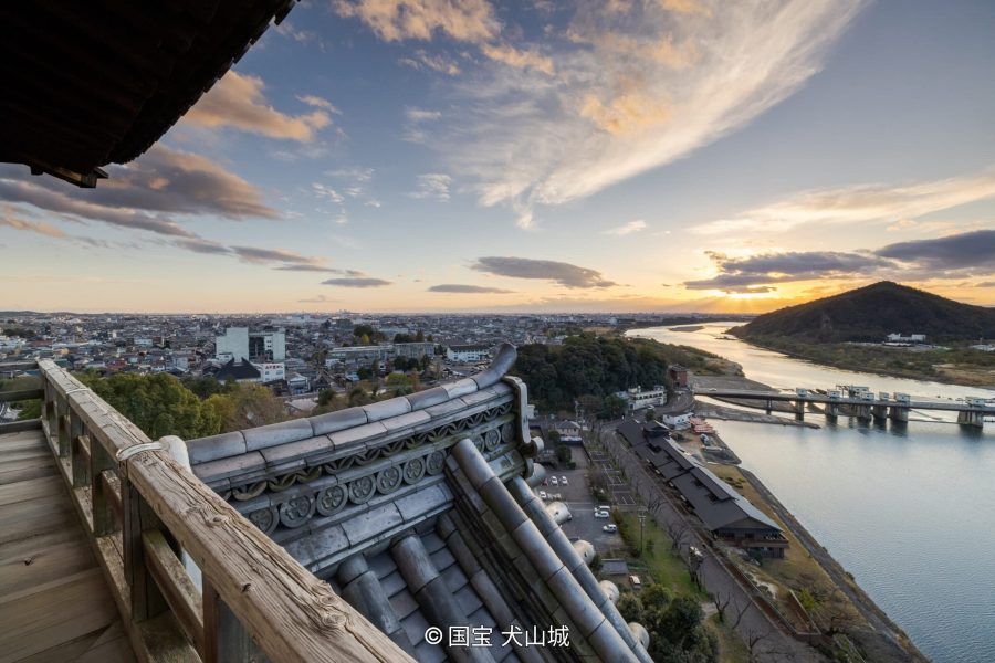 A panoramic view of the city, the Kiso River, and a mountain taken from the top level of Inuyama Castle at sunset, showing the castle's tiled roof and wooden railing in the foreground.