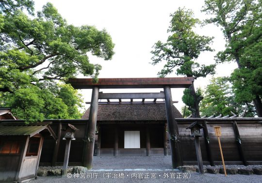 A massive wooden torii gate marking an inner entrance (likely to the Naiku or Geku) of Ise Jingu Shrine, with a traditional fence and thatched roof structure behind it.