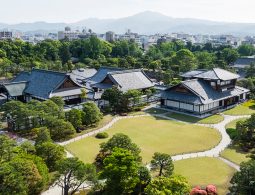 An aerial view of the traditional architecture of Nijo-jo Castle's Ninomaru Palace and the sprawling, manicured gardens, with the city of Kyoto in the background.