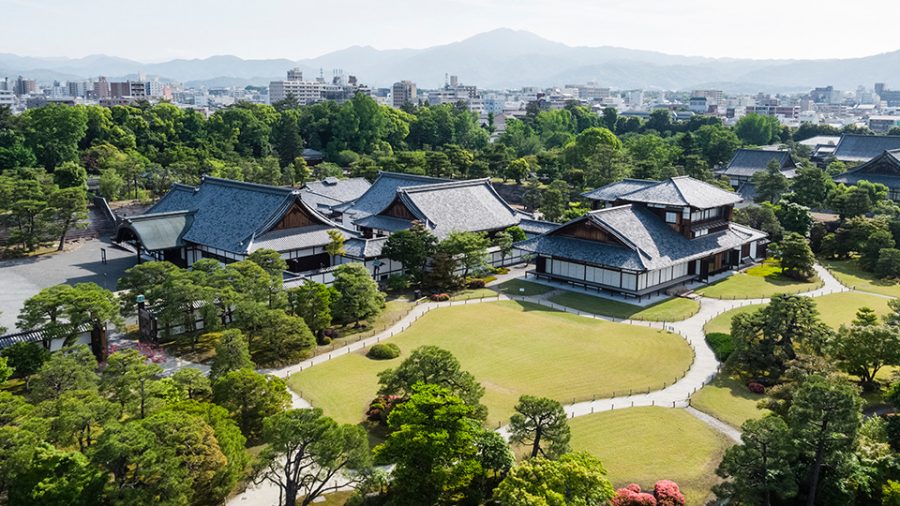 An aerial view of the traditional architecture of Nijo-jo Castle's Ninomaru Palace and the sprawling, manicured gardens, with the city of Kyoto in the background.
