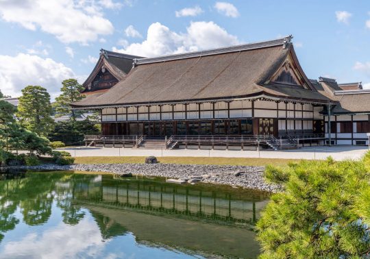 The Oikeniwa Garden at Kyoto Imperial Palace, viewed over the pond with reflections of the traditional building and surrounding pine trees.