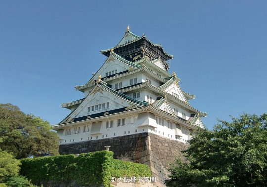 The majestic white and green-tiled main tower of Osaka Castle, sitting atop massive stone walls under a clear blue sky.
