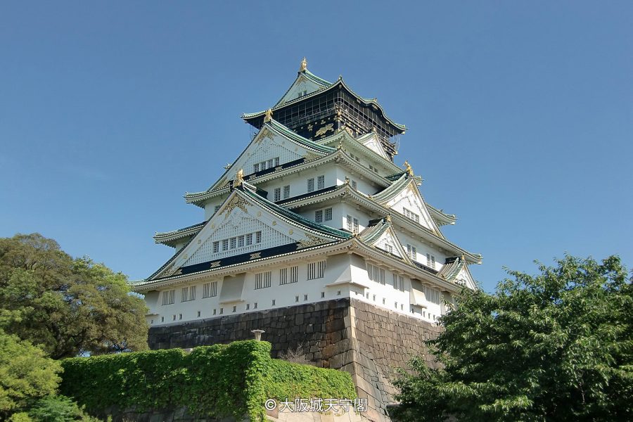 The majestic white and green-tiled main tower of Osaka Castle, sitting atop massive stone walls under a clear blue sky.