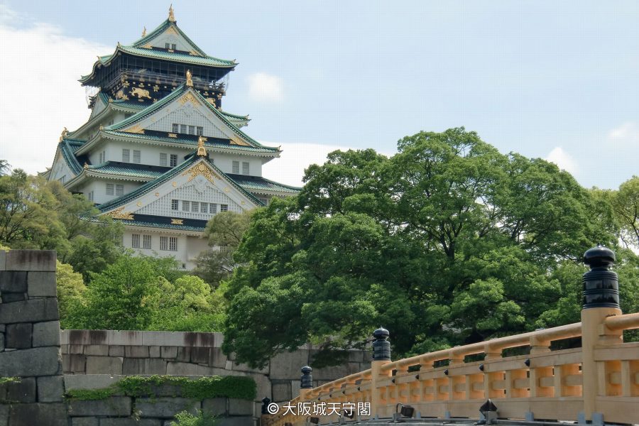 The main keep of Osaka Castle visible beyond a curved wooden bridge and lush green trees in the foreground.