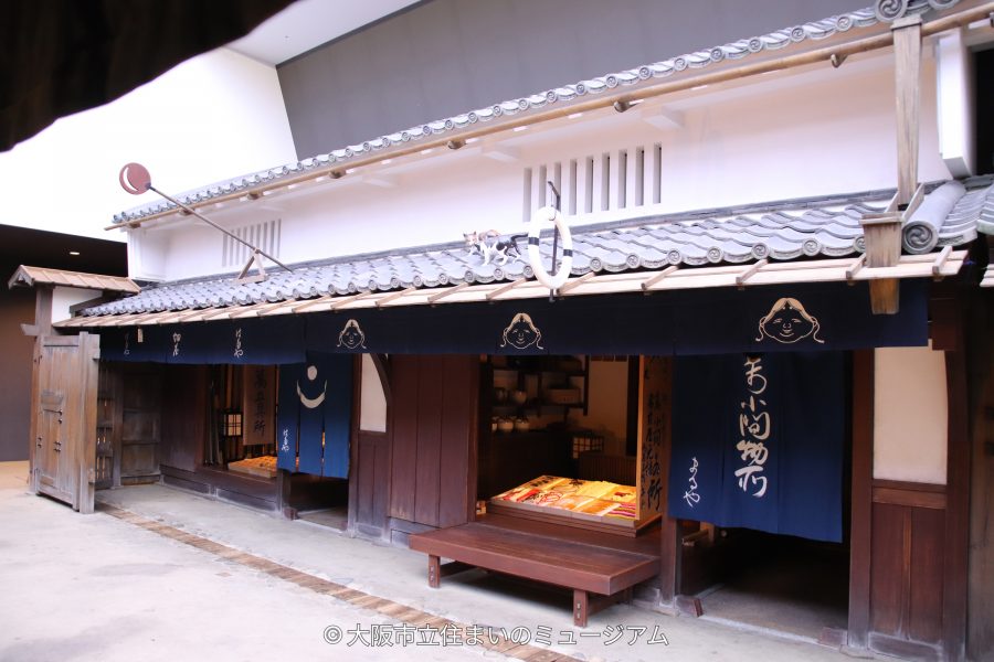 The facade of a recreated Edo period shop (likely a small merchant or general store) with noren curtains and a low counter.