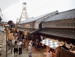 A bustling, high-angle view of the recreated Edo period Osaka streetscape, showing visitors and paper lanterns below a tall wooden fire tower.