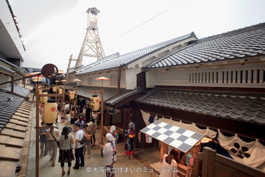 A bustling, high-angle view of the recreated Edo period Osaka streetscape, showing visitors and paper lanterns below a tall wooden fire tower.