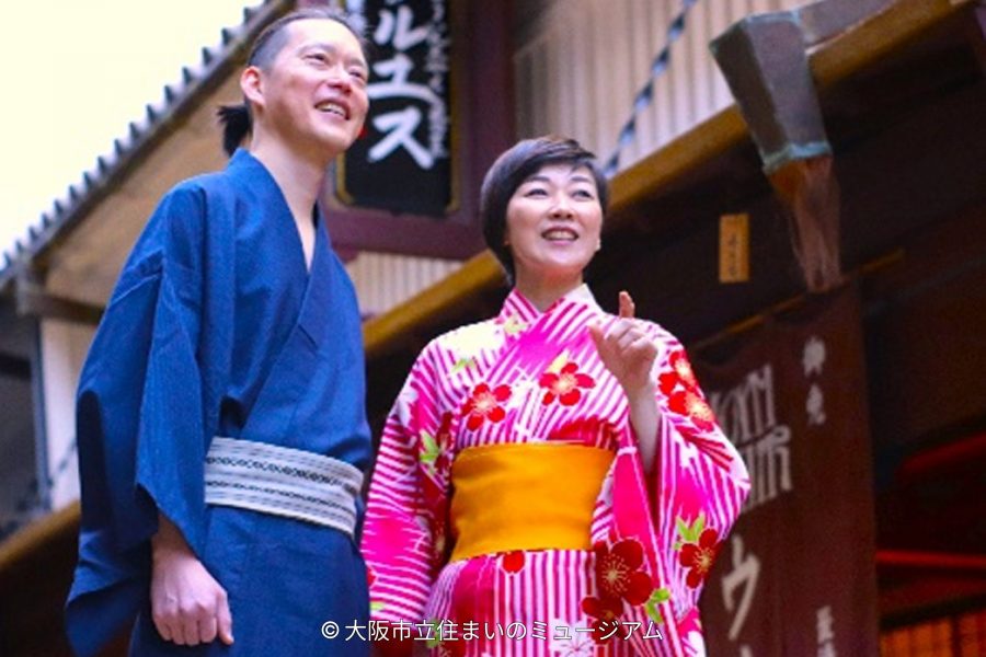 A smiling man in a blue yukata and a woman in a bright pink floral yukata posing in a recreated Edo period street setting.